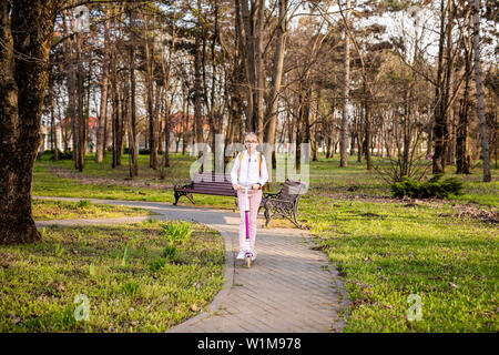 Lustige blonde Mädchen lernen auf Weiß scooter im warmen grünen Park in der Mitte der großen Stadt zu skaten. Sommer aktivitäten Konzept. Stockfoto