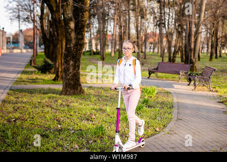 Lustige blonde Mädchen lernen auf Weiß scooter im warmen grünen Park in der Mitte der großen Stadt zu skaten. Sommer aktivitäten Konzept. Stockfoto