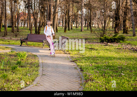 Lustige blonde Mädchen lernen auf Weiß scooter im warmen grünen Park in der Mitte der großen Stadt zu skaten. Sommer aktivitäten Konzept. Stockfoto