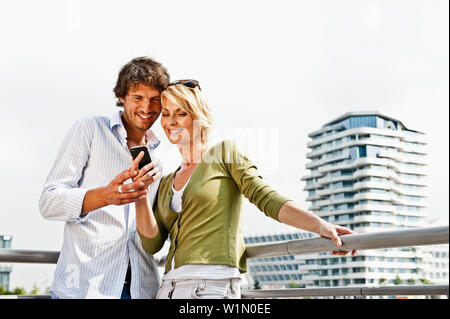 Lächelndes Paar mit Handy lehnte sich gegen Balustrade, Marco-Polo-Tower im Hintergrund, HafenCity, Hamburg, Deutschland Stockfoto
