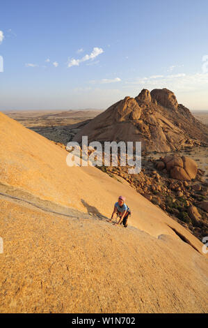 Frau Klettern am roten Fels, Sugarloaf im Hintergrund, Große Spitzkoppe, Namibia Stockfoto