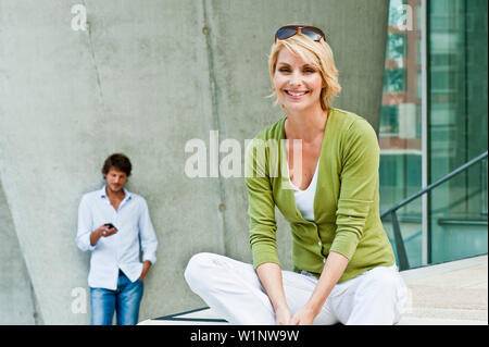 Frau sitzt auf Schritt, Mann mit Handy stehen im Hintergrund, HafenCity, Hamburg, Deutschland Stockfoto