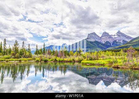 Die Canmore berühmten Three Sisters Berggipfel und deren Reflexionen auf dem Bow River im südlichen Banff Spektrum der Kanadischen Rockies an einem bewölkten S Stockfoto