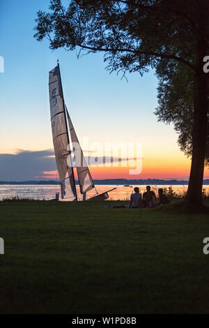 Katamaran ist im letzten Abendlicht am Chiemsee Ufer, sitzen drei Jugendliche unter einem Baum Stockfoto