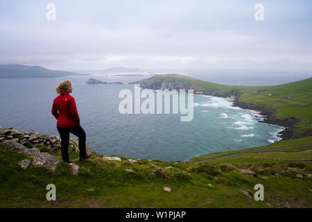 Eine blonde Frau in einem roten Mantel und Wanderschuhe genießt die Aussicht von Slea Head und der schlafende Riese der großen blaskets an einem windigen Frühlingstag gesehen fr Stockfoto