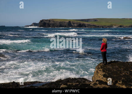 Wellen des Atlantischen Ozeans Crash gegen die Felsen, wo eine Frau in einem roten Mantel Uhren aus entlang der Steilküste Kilkee gehen, während ihr Haar aro Schlagsahne bekommt Stockfoto