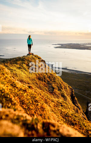 Junge Frau auf einem moosy Felsen hoch über dem Meer, Sonnenuntergang, Johannesburg, Vesturland, Island Stockfoto