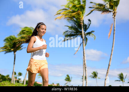 Asiatische runner girl Jogging in tropischen Natur Park im Freien mit Palmen Hintergrund im Sommer. Junge Erwachsene sport Frau Laufen fit tragen orange Fitness Shorts und Tank Top. Stockfoto