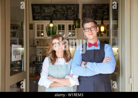 Inhaber kleiner Unternehmen. Schuß von Frau mittleren Alters und jungen Mann mit Schürze und stehen mit den Armen in unserem kleinen Cafe gekreuzt, während für das Warten Stockfoto
