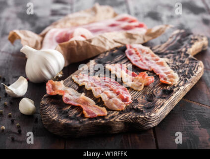 Gegrilltem Speck Streifen auf vintage Holzbrett mit rohen frisch geräuchertem Schweinefleisch Speck auf Metzger Papier auf Holz mit Knoblauch. Stockfoto