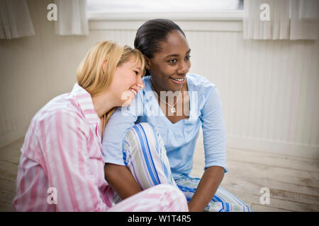 Zwei lächelnde junge Frau, die neben einander sitzen auf dem Boden tragen Schlafanzug. Stockfoto
