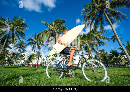 Surfer mit blonden Haaren mit seinem Surfbrett auf einem Beach Cruiser Fahrrad durch eine tropische Palme Landschaft in Miami, Florida, USA Stockfoto