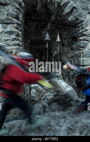 Zwei junge Skifahrer zu Fuß vorbei an kleinen Glocken einer Ruine, Gudauri, Mtskheta-Mtianeti, Georgien Stockfoto