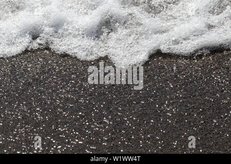 Wave Schaum auf dem Sand. Nahaufnahme des Meeres Wellen mit weißen Schaum auf dem sandigen Ufer mit Sun Reflexionen Wirkung. Stockfoto