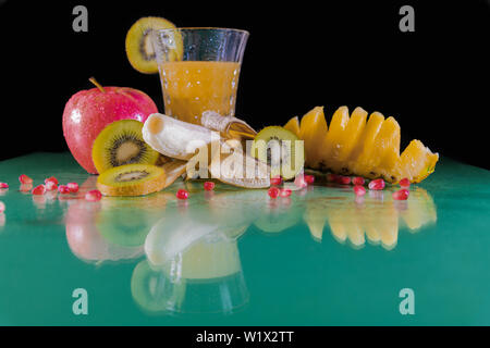 Nahrhafte Banane, in Scheiben geschnittene grüne Kiwi, Apfel, Hawaiianische Ananas- und Orangensaft auf einem grünen Tisch - Schwarzer Hintergrund-Close-up Stockfoto