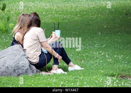 Zwei Mädchen sitzen auf einem grünen Gras, trinken Cocktails und Eindrücke auszutauschen. Sommer Freizeitaktivitäten auf ein Kleeblatt Wiese, Konzept der weiblichen Freundschaft Stockfoto