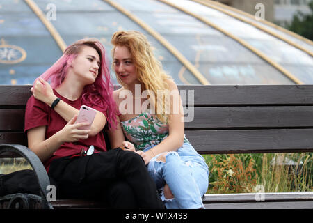 Zwei hübsche Mädchen sprechen auf einer Bank auf Manezh Square, jugendlich mit rosa Haaren hält Smartphone. Konzept der weiblichen Freundschaft, Gefühle, Klatsch Stockfoto