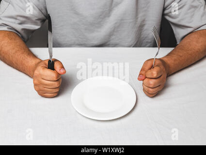 Mann sitzt hinter einem Tisch mit Messer und Gabel in den Händen und leeren Teller, Zeit zu essen - heilfasten Konzept Stockfoto