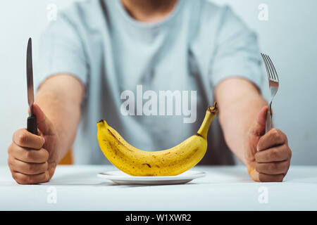 Mann hinter einem Tisch mit Messer und Gabel in den Händen und einem frischen Bananen sitzen auf einer Platte auf einem weißen Tisch, Zeit zu essen - gesunde ernährung Konzept Stockfoto