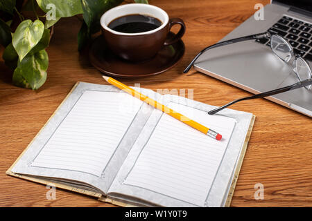 Leer Notebook und pencill auf einem Schreibtisch mit Tasse Kaffee und Laptop im Hintergrund geöffnet