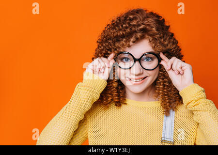 Curly - behaarte Frau ihre Brille ajusting auf orange Hintergrund. tennage Mädchen mit stilvollen hipster Gläser Stockfoto