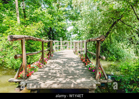 Hand Made romantische kleine Holzbrücke über den Fluss von Wasser durch die Bäume und Blumen, die zu Pier unter Fluss Pflanzen im Alter von verwittertes Holz mad umgeben Stockfoto