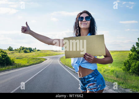 Close up Girl reist per Anhalter mit einem Karton Zeichen in den Händen. Platz für Text. Ein Mädchen in Gläser Shorts und Heels Straße am Straßenrand halten einer em Stockfoto