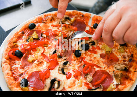 Des Menschen Hand schneiden Salami Pizza mit machrooms, Oliven, Käse. Stockfoto