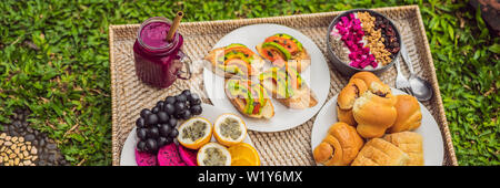 BANNER, LANGE FORMAT Frühstück auf einem Tablett mit Obst, Brötchen, Avocado Sandwiches, smoothie Schüssel stehen auf dem Gras Stockfoto