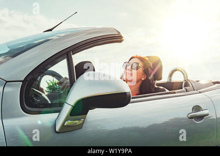 Sommer road trip. Sie reist nach einem Gig mit offenem Dach. Zufriedene Frau sitzt entspannt im Auto gegen den Himmel mit Sonne. Das Konzept der trav Stockfoto