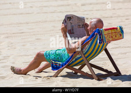 Mann sitzt im Liegestuhl Lesen der Zeitung am Strand von Bournemouth, Dorset Großbritannien im Juli Stockfoto