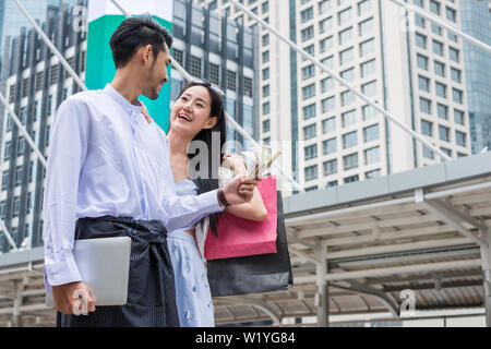 Geschäftsmann holding Dollar cash mit Laptop und asiatische Frau mit einkaufskorb sind glücklich lächelnd in die Stadt Stockfoto