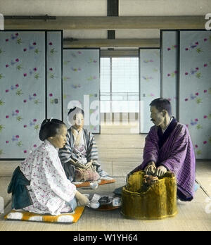 [1930er Jahre Japan - Tee] - zwei Frauen und ein Mann in traditioneller Kleidung sitzen auf ZABUTON (Kissen) auf Tatami Matten in einem großen Zimmer geteilt durch Fusuma (Schiebetüren) an, was scheint, eine Inn zu sein. Der Mann ist Erwärmung seine Hände an einem schönen hölzernen Hibachi für das Halten das Wasser warm verwendet. Die Frau in der vorderen erscheint Tee zu bereiten. 20. Jahrhundert vintage Glas schieben. Stockfoto