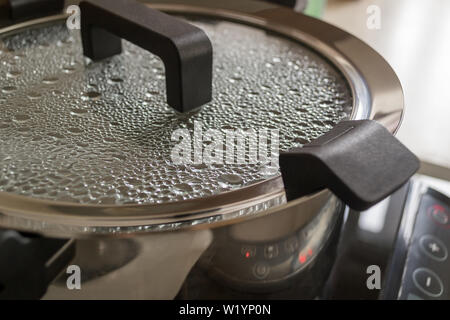 Modernes Metall Suppe Topf oder Pfanne mit schwarzen Griffen auf einem Induktionsherd. Tropfen von kochendem Wasser sind auf der Innenseite des durchsichtigen Glas Deckel. Stockfoto