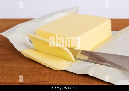 Ein paar Scheiben gelb Butter abgeschnitten von einer gerade eröffnet Pack von Butter mit einem Messer auf einem braunen Holz Schneidebrett. Stockfoto