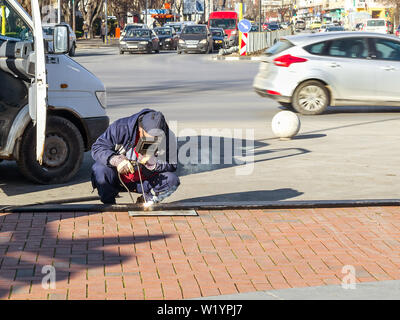 Ein Schweißer mit Gesichtsschutz und Handschuhe hockt und führt eine Schweißarbeiten oder dringende Reparaturen auf einer Straße der Stadt an einem sonnigen Tag. Stockfoto