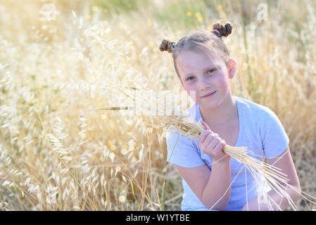 Schöne Mädchen, dass Spikes von Weizen und Hafer. Niedliche Kind sitzen auf den goldenen Herbst Feld zur Ernte bereit. Stockfoto