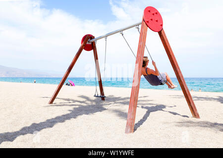 Frau mittleren Alters Schwingen auf einer Schaukel. Am Strand mit der Sonne auf Ihrem Gesicht in Spanien. Stockfoto