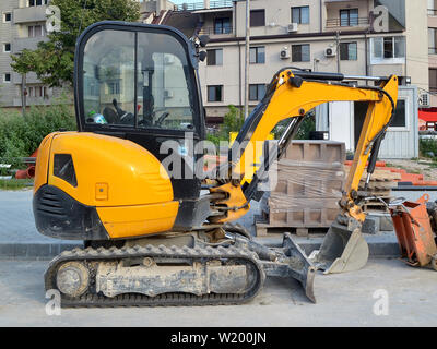 Gelbe Mini Bagger an den Anschlüssen für kleine Bauarbeiten an schwer zugänglichen Stellen oder auf schmalen Straßen der Stadt, Seitenansicht, Sommertag. Stockfoto