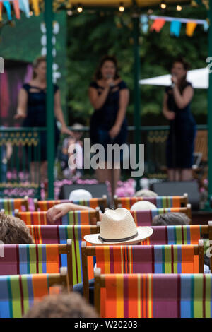 Die Menschen saßen in Liegestühlen beobachten eine weibliche Vintage boogie - woogie Gesangsgruppe im musikpavillon an der RHS Hampton Court Flower Show 2019. Surrey, Großbritannien Stockfoto