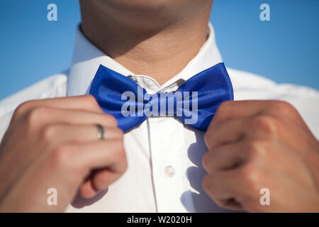 Ein Bräutigam in einem weißen Hemd richtet eine blaue Schleife vor blauem Himmel Stockfoto