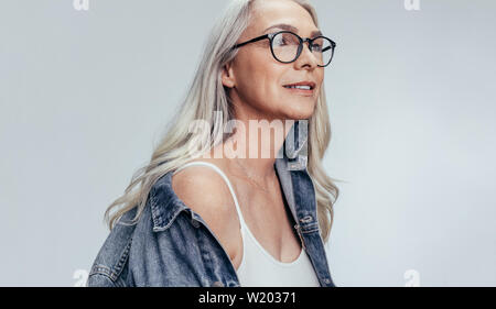 Schöne ältere Frau in Casuals bei copy Raum gegen grauen Hintergrund. Stilvolle Mitte nach kaukasische Frau in Denim Shirt und Brille looki Stockfoto