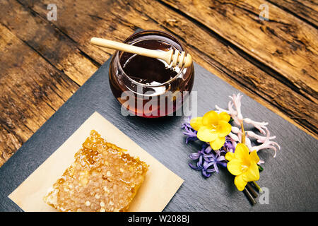 Gesunde, natürliche, organische und süßen Honig im Glas oder Schüssel mit Honig Pendelarm, Blumen und Wabe auf schwarzem Hintergrund. Stockfoto