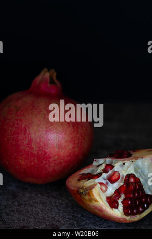 Ein ganzes Granatapfel (Punica Granatum) und ein Schnitt mit leuchtend roten Samen auf eine strukturierte graue Oberfläche vor einem schwarzen Hintergrund Stockfoto