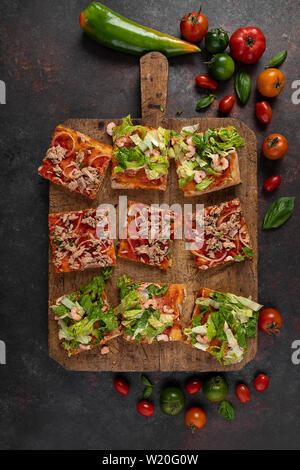 Pizza Scheiben mit Garnelen, Salat, Tomaten, Thunfisch und Zwiebeln, auf einer hölzernen Küche board, Ansicht von oben geschossen. Stockfoto