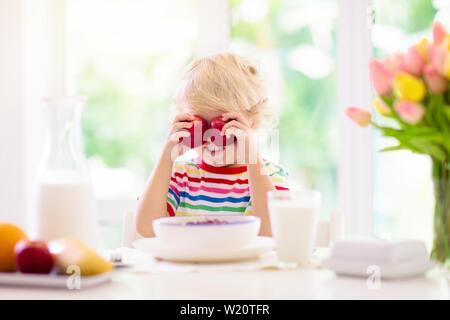 Kind frühstücken. Kind Essen trinken Milch und Müsli mit Obst. Kleiner Junge am weißen Esstisch in der Küche am Fenster. Kinder Essen an sonnigen morni Stockfoto