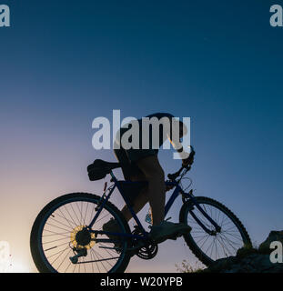 Silhouette eines passenden männlichen Mountain Biker, sein Fahrrad bergauf auf felsigen rauen Gelände auf einem Sonnenuntergang. Stockfoto