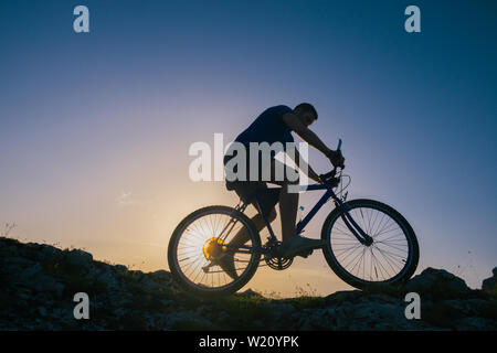 Silhouette eines passenden männlichen Mountain Biker, sein Fahrrad bergauf auf felsigen rauen Gelände auf einem Sonnenuntergang. Stockfoto