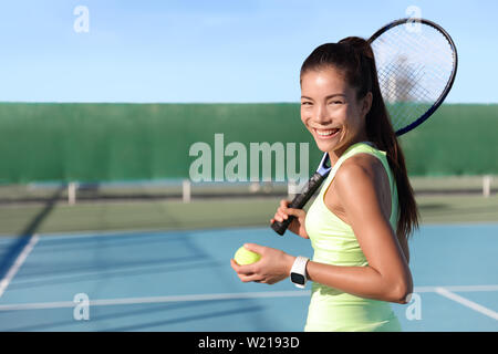 Asiatische junge Tennisspieler Frau auf outdoor Hard Court holding Ball und Schläger portrait. Weibliche Athleten tragen gelbe Sportbekleidung und Activity Tracker smartwatch. Wearable tech fitness Watch. Stockfoto