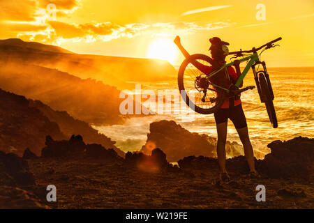 Erfolg, Verwirklichung, Vollendung und gewinnende Konzept mit Radfahrer Mountainbiken. Gerne MTB Frau radfahren Anheben der Arme heben Bike vom Meer bei Sonnenuntergang Jubeln und Feiern auf dem Gipfel. Stockfoto